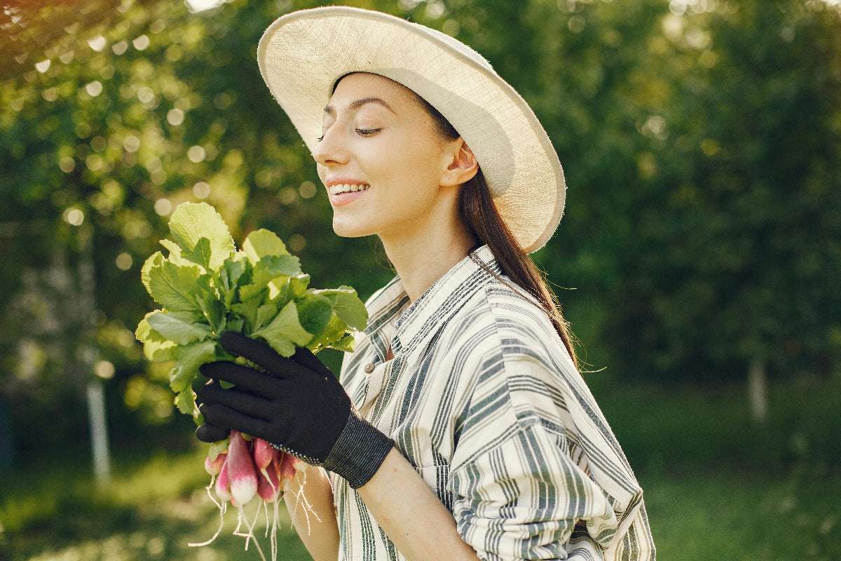 Frau mit Hut und Handschuhen steht im Garten und freut sich über frisch geerntete Radieschen