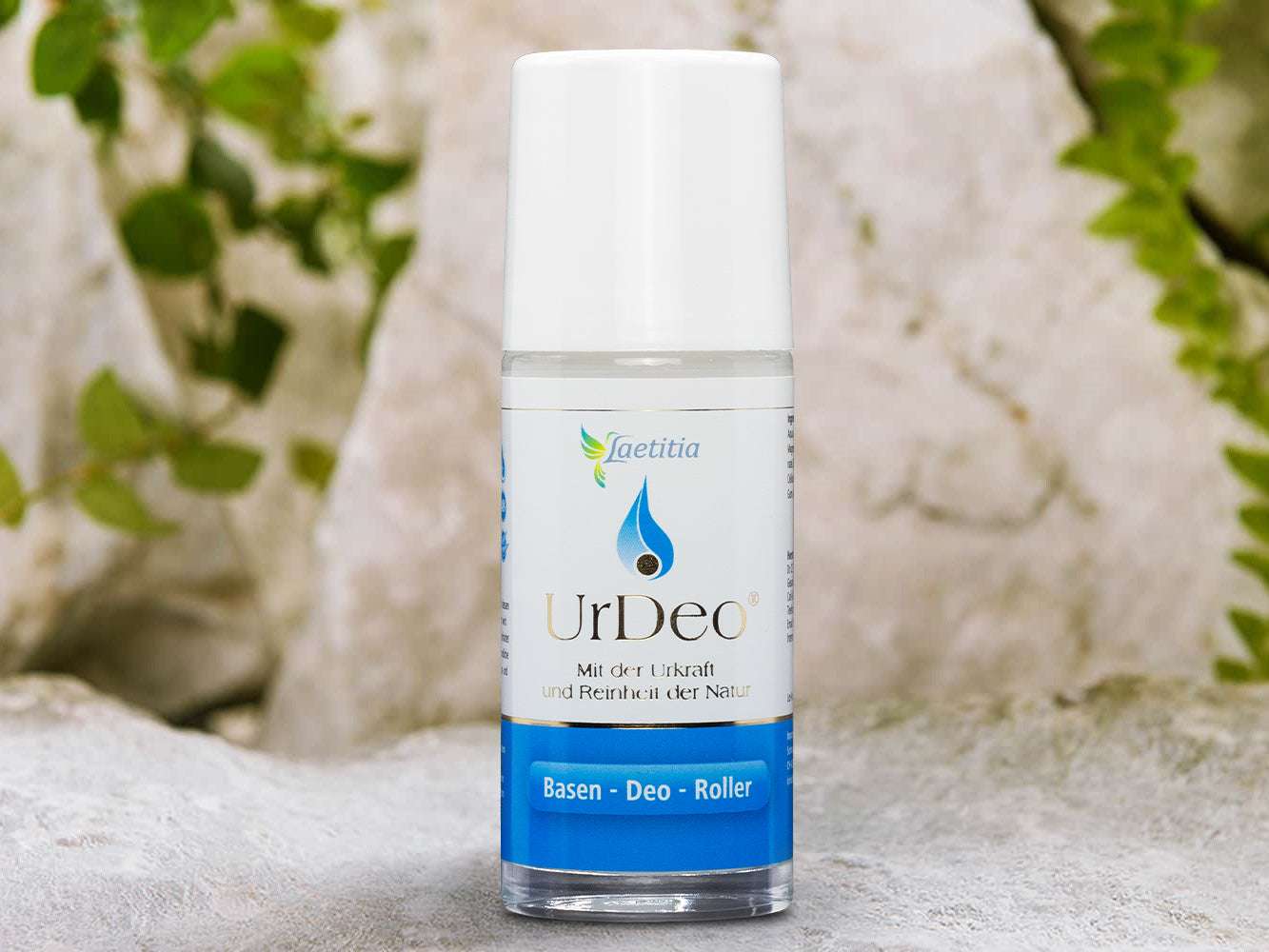 Basisches UrDeo von Laetitia vor einem natürlichen Hintergrund aus Felsen und Pflanzen, blau-weiße Verpackung, Roll-on-Flasche.