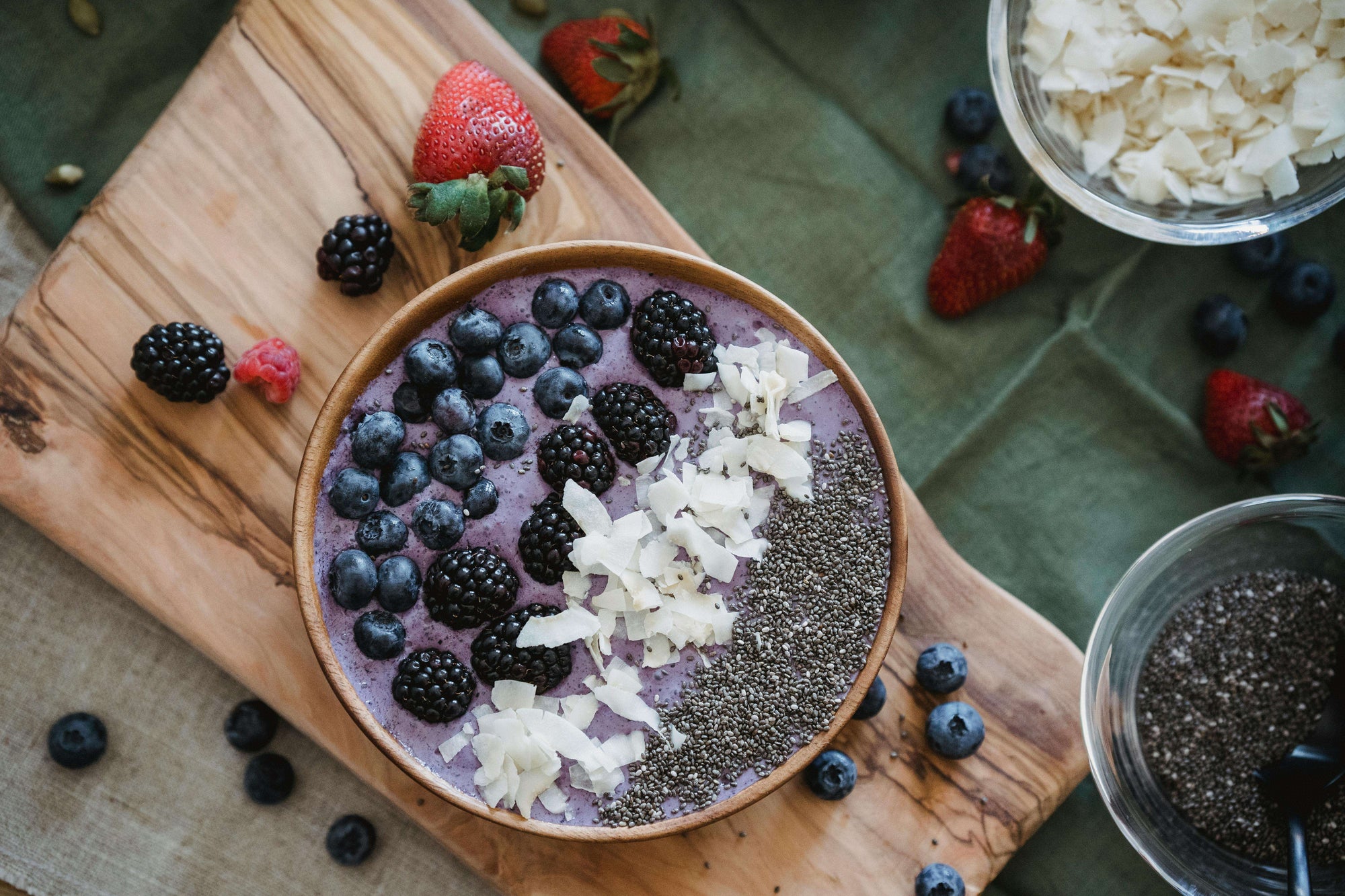 Schale mit Joghurt, Blaubeeren, Brombeeren, Kokosflocken und Chia Samen auf einem Holzbrett. Außen herum noch Himbeeren und Erdbeeren als Dekoration
