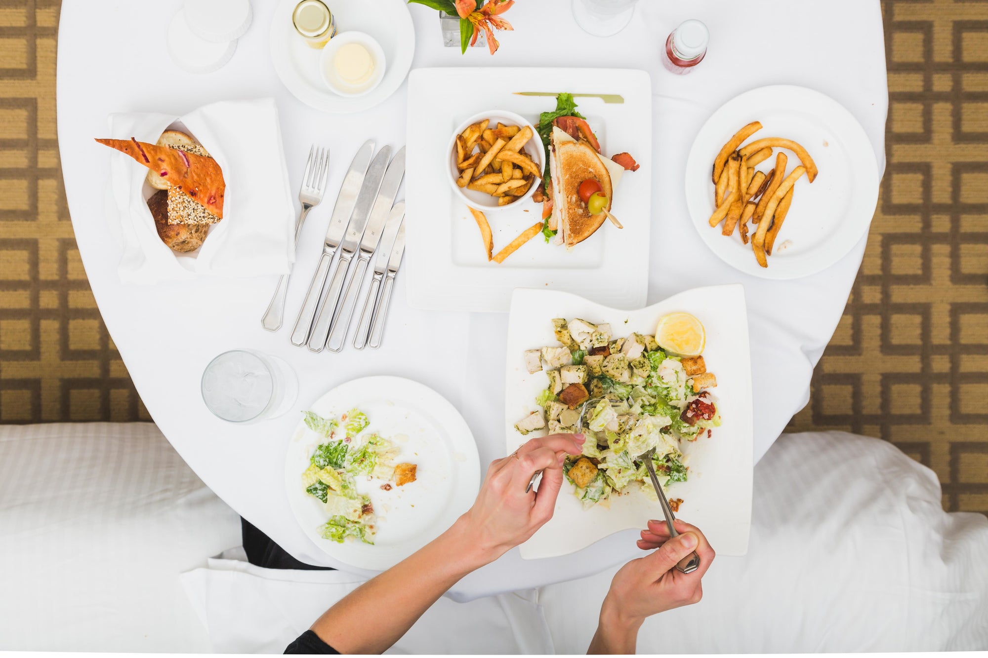 Gedeckter Tisch mit verschiedenen Speisen: ein frischer Caesar-Salat mit Croutons, Hähnchen und Zitronenscheibe, ein Sandwich mit Pommes frites, ein Brotkorb sowie leere und halbvolle Teller. Zwei Hände greifen zur Gabel – Szene eines Hotel-Frühstücks oder Room-Service-Mahls aus der Vogelperspektive.