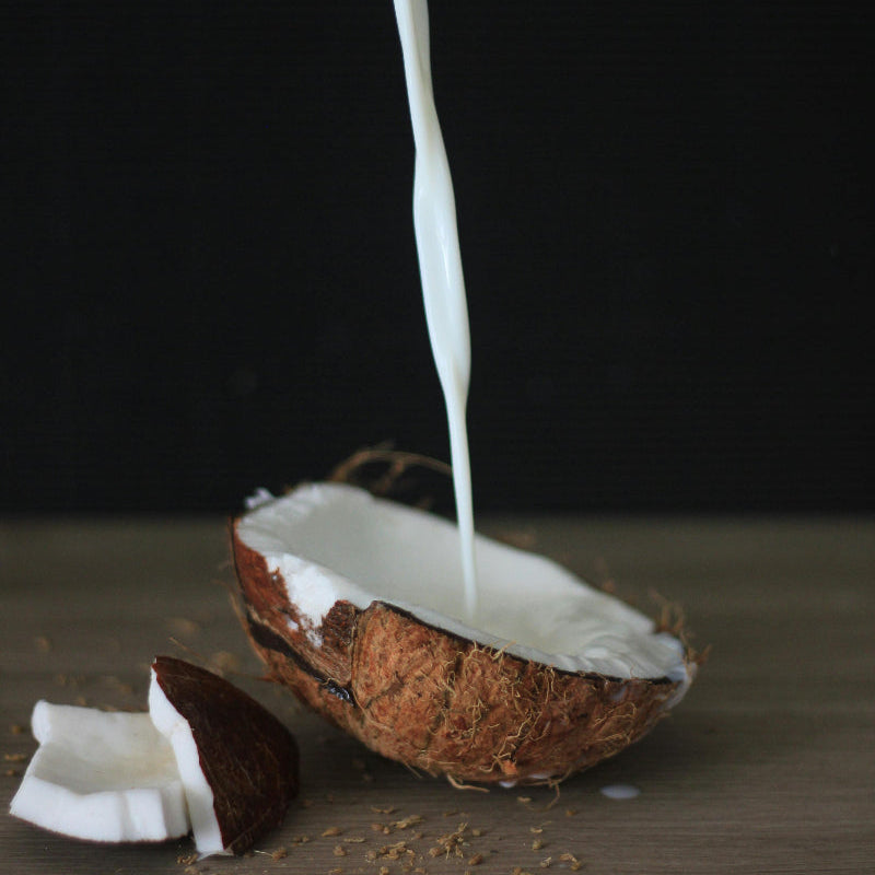 Ein Strahl Kokosmilch wird in eine halbierte Kokosnuss gegossen, die auf einer Holzoberfläche liegt. Daneben liegen Stücke von Kokosnussfleisch. Das Bild veranschaulicht Kokosmilch als pflanzliche Milchalternative.