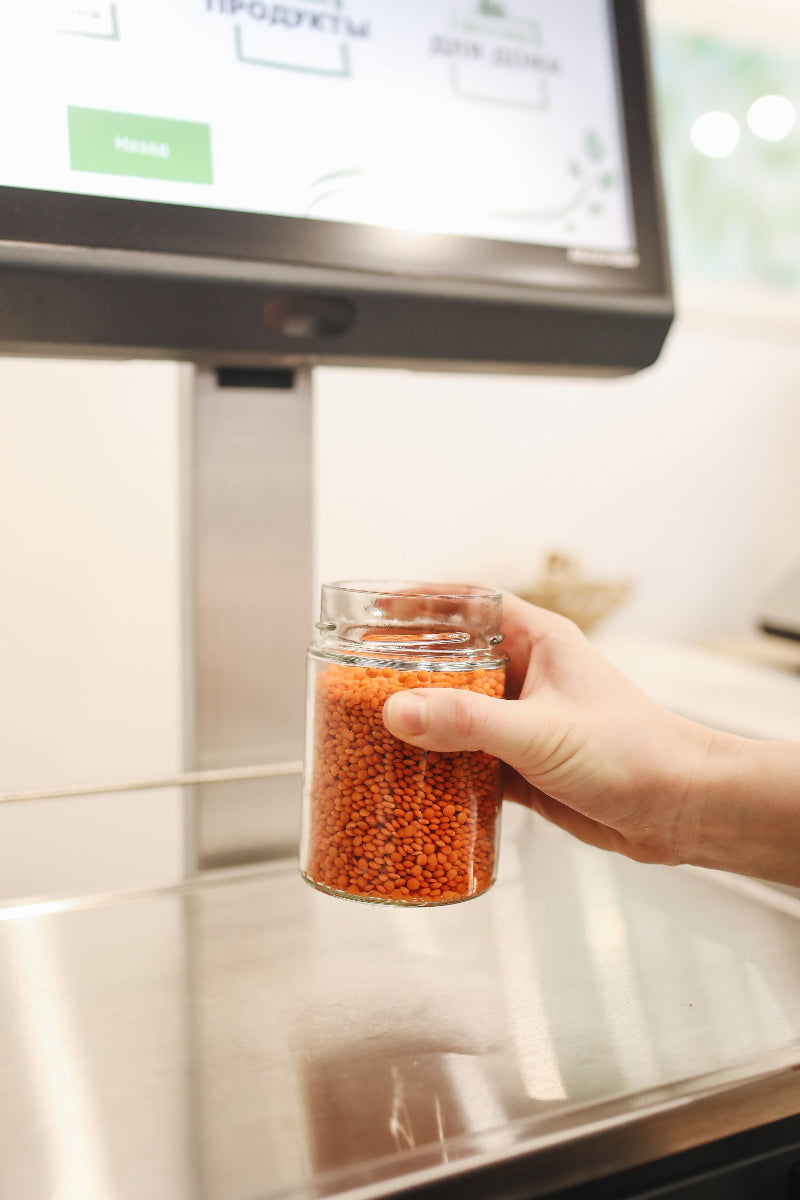 Hand hält ein Glas mit roten Linsen vor einer Waage oder Analyse-Station. Das Bild begleitet einen Beitrag zur Frage, ob Linsen basisch sind und wie sie im Säure-Basen-Haushalt eingeordnet werden.