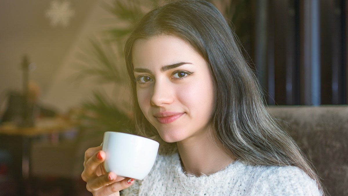 Junge Frau trinkt Kaffee aus einer weißen Tasse in einem Wohnzimmer mit einer Pflanze im Hintergrund