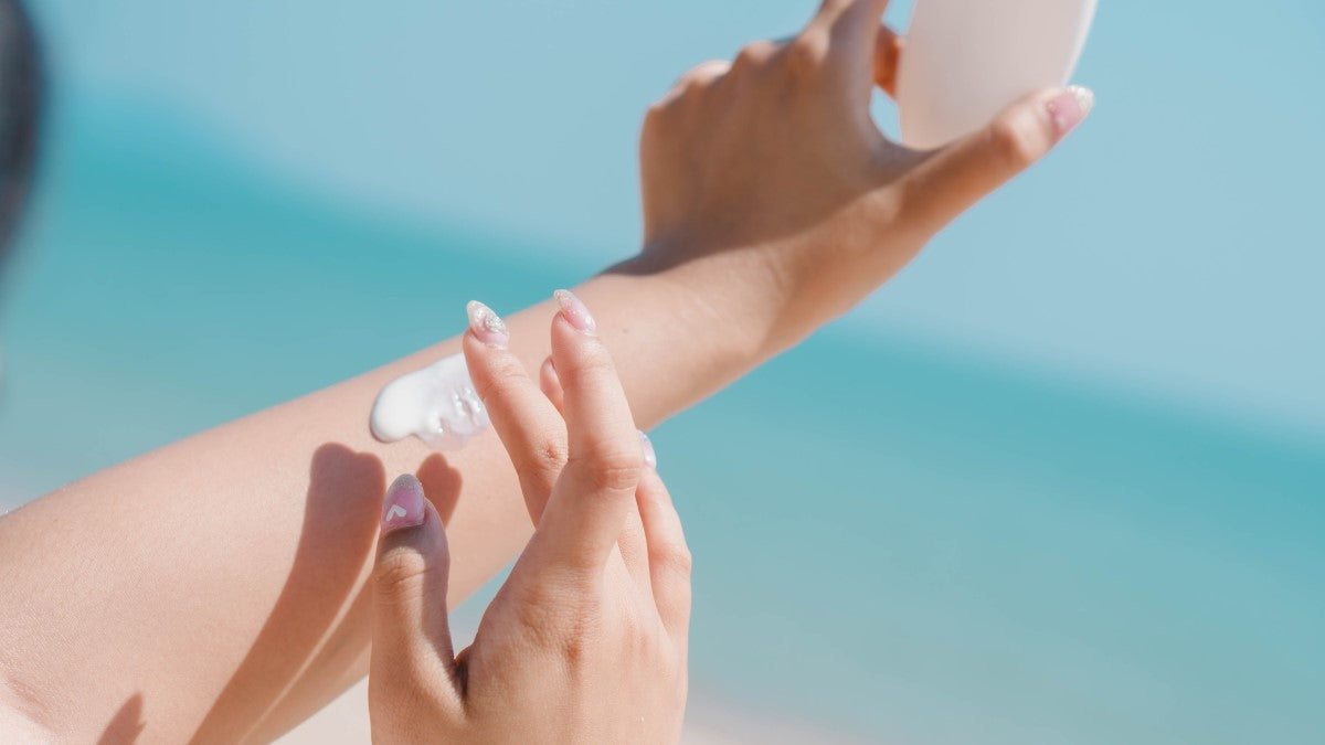 Person trägt Sonnencreme auf den Arm auf, im Hintergrund Meer und blauer Himmel; Symbolbild für Sonnenschutz und Sommerreiseapotheke mit Sonnencreme