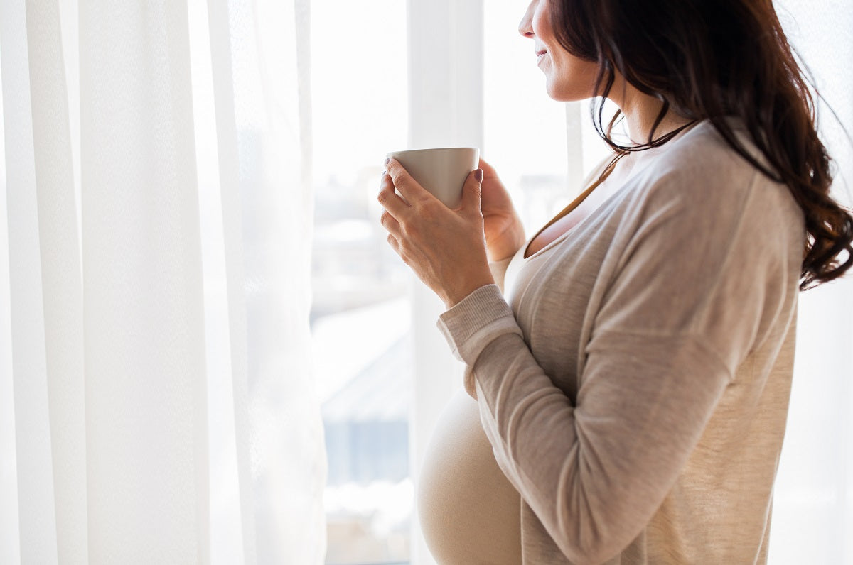 Schwangere Frau in hellen Klamotten genießt lächelnd eine Tasse Tee vor dem Fenster